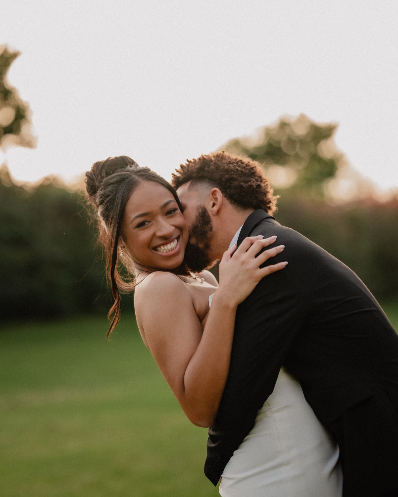 Bride laughing wide-open as her groom kisses her cheek in golden field light