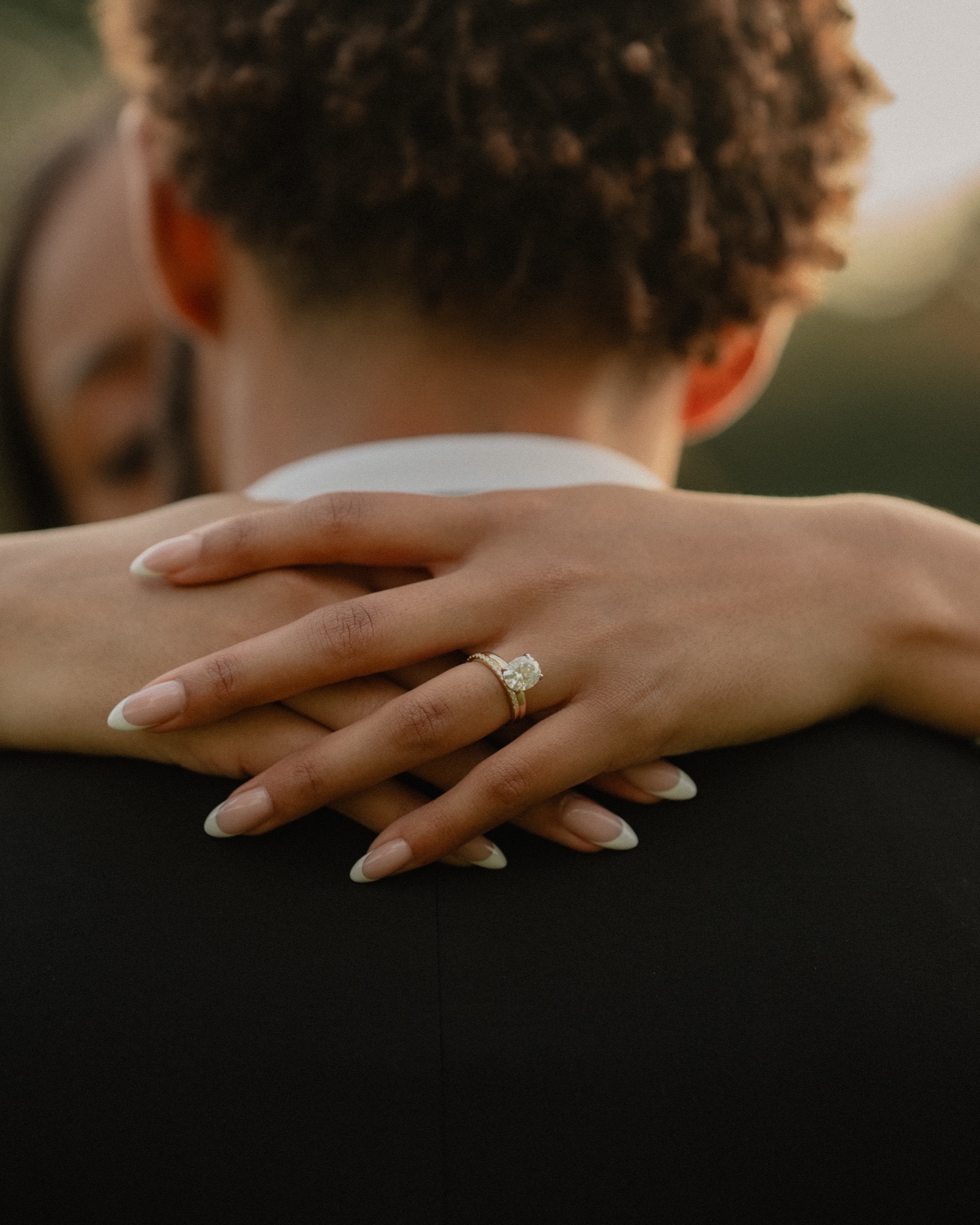 Bride's hands wrapped around her groom's shoulder, engagement ring catching the warm afternoon light