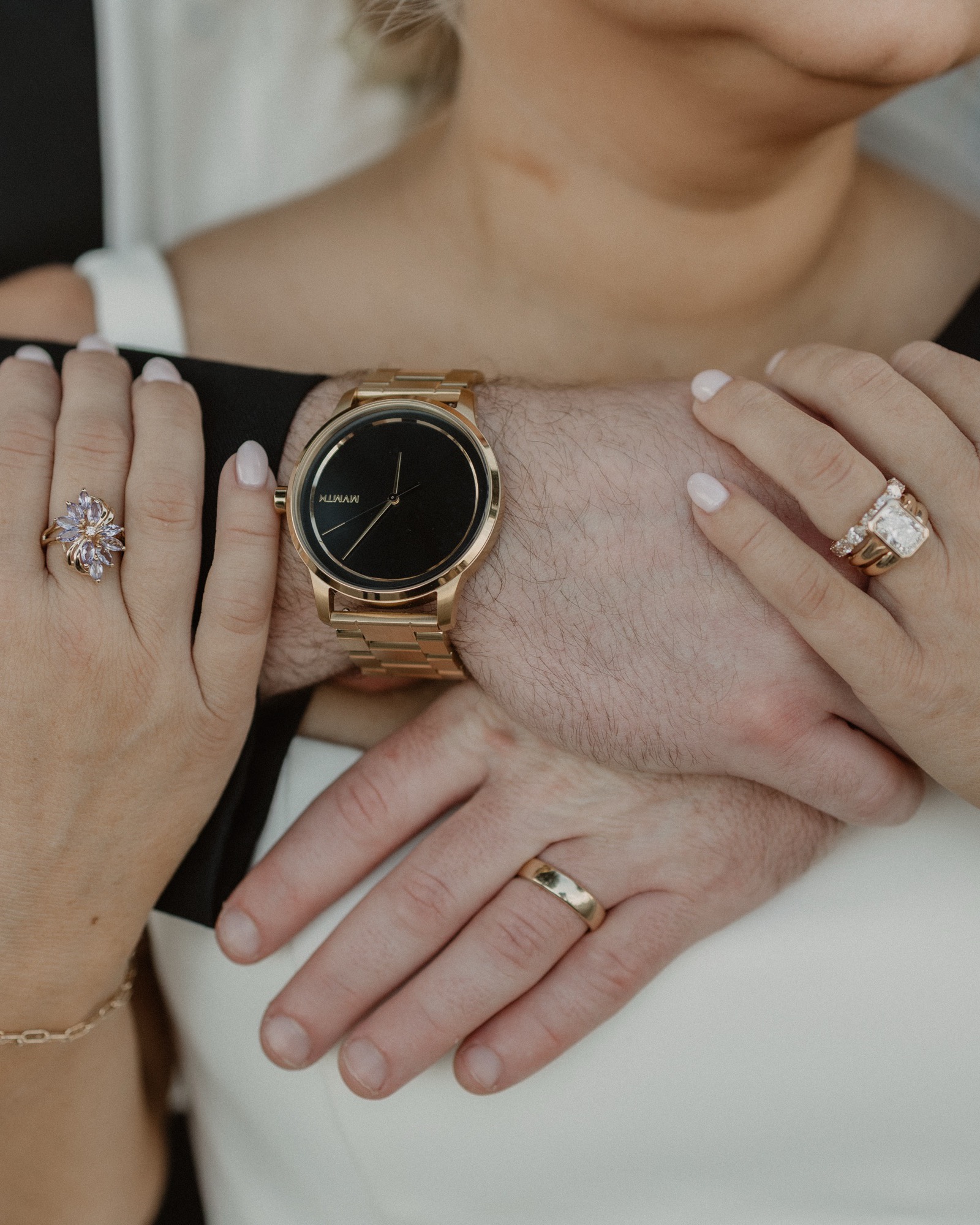Bride and groom hands intertwined, gold watch and wedding rings centered in the frame