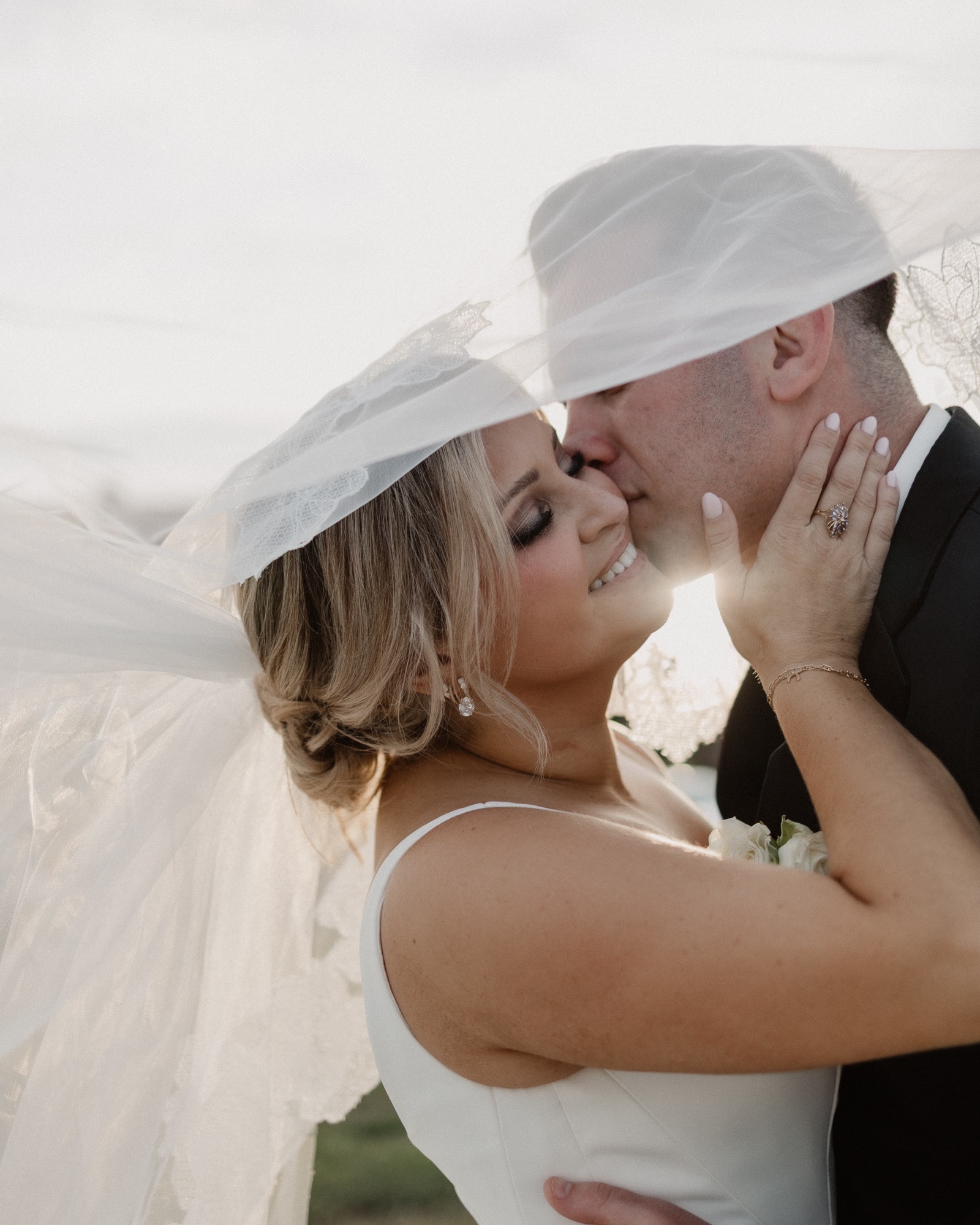 Bride and groom kiss beneath a flowing veil, sun bursting behind them at golden hour